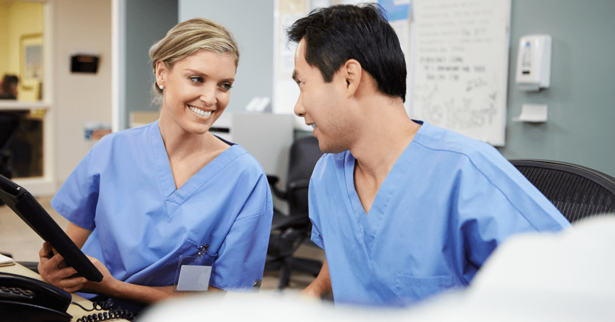 Two nurses in blue scrubs conferring at a hospital unit workstation