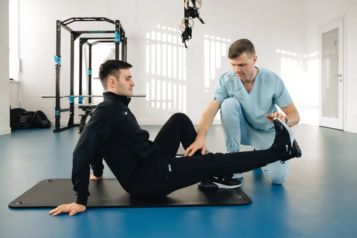 A physiotherapist in blue scrubs assisting a man with a leg exercise on a mat in a rehabilitation clinic.