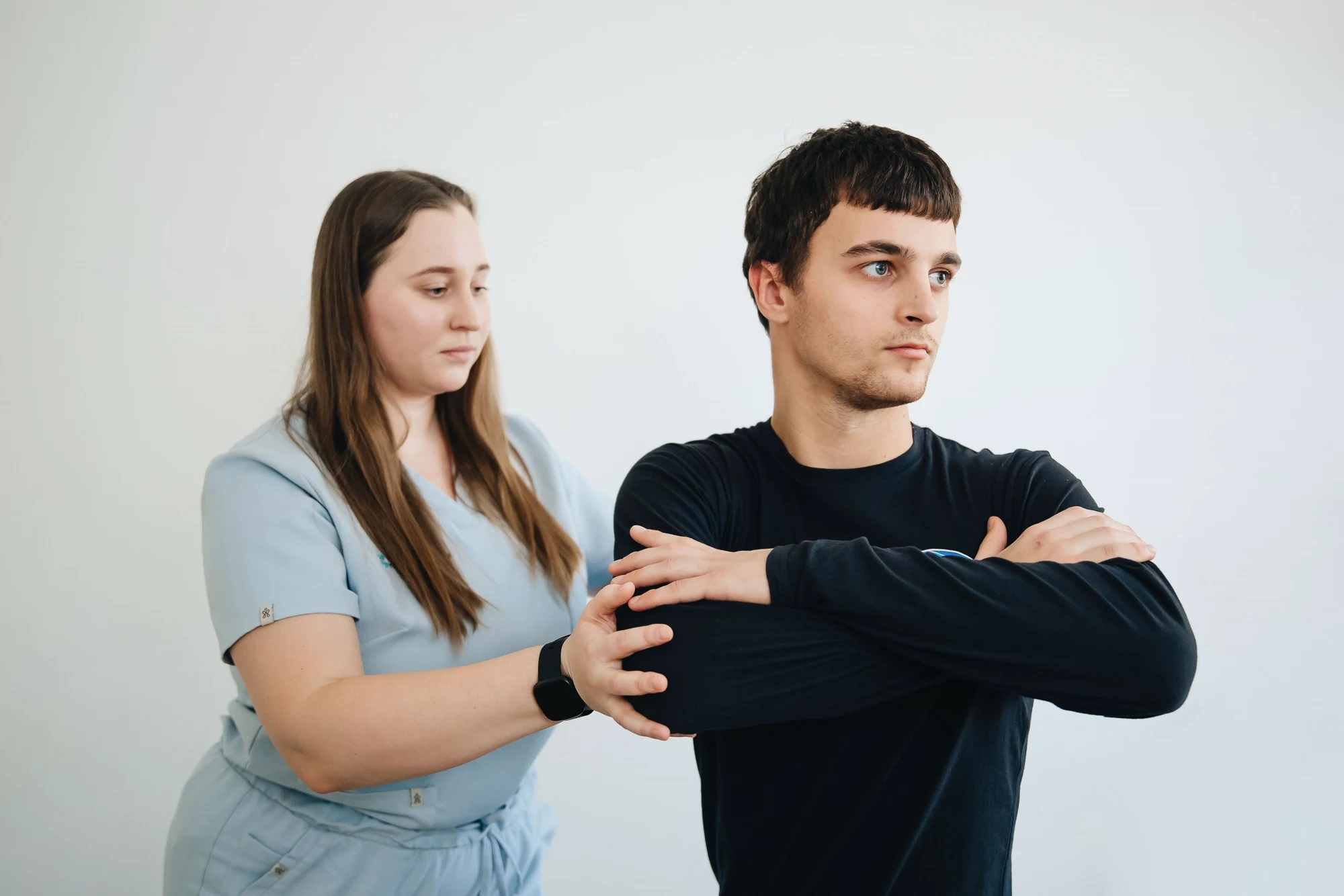 A physiotherapist in blue scrubs guiding a seated man through an arm and shoulder exercise.