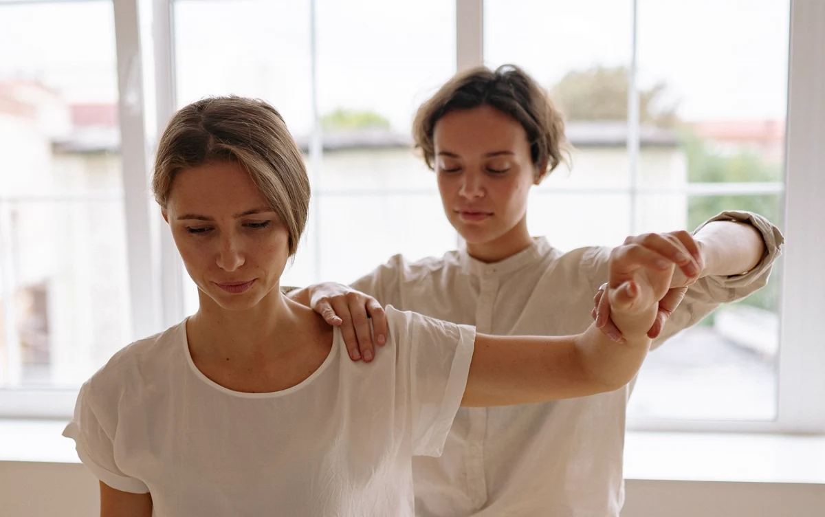 A therapist guiding a seated woman through a raised arm exercise beside a treatment table.