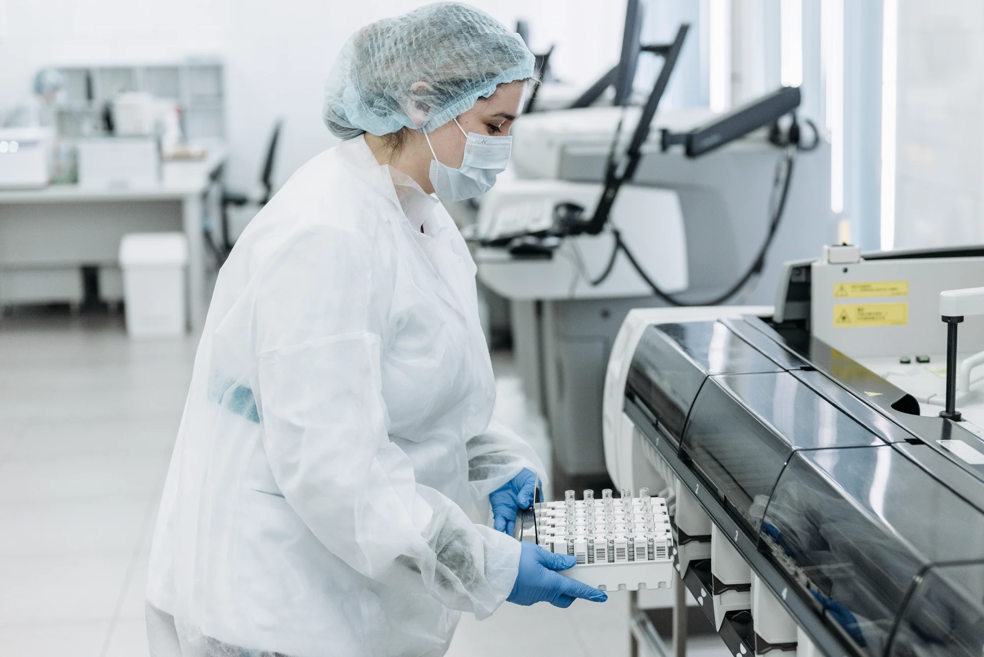 A laboratory professional in protective clothing and blue gloves holding a rack of sample tubes beside an analyser.