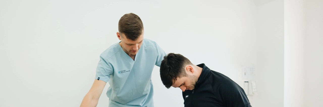 A physiotherapist in blue scrubs guiding a seated man through a rehabilitation exercise.